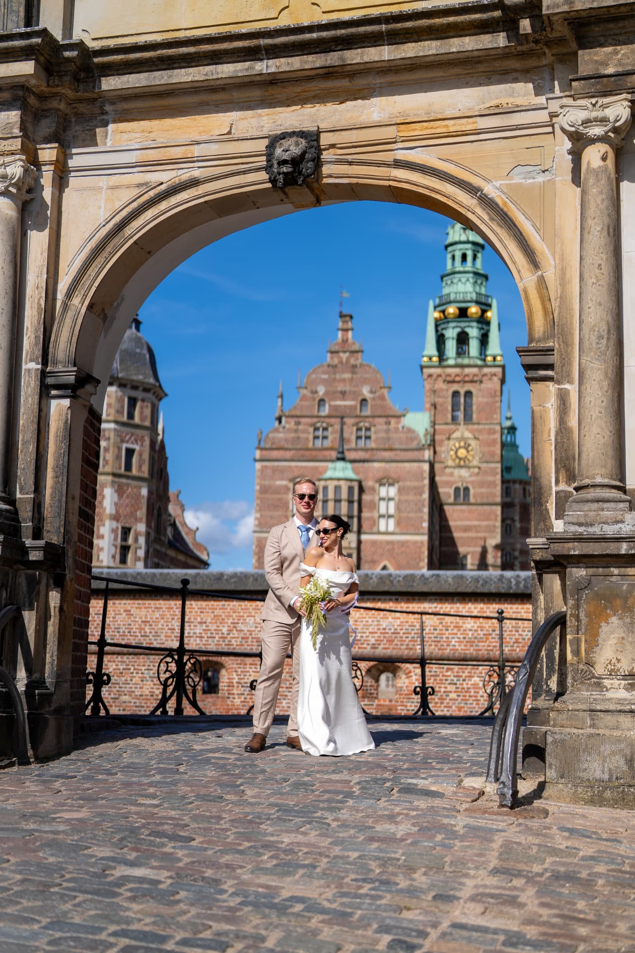 Castle wedding photography Frederiksborg Denmark — bride and groom framed by the stone archway with the castle towers behind them