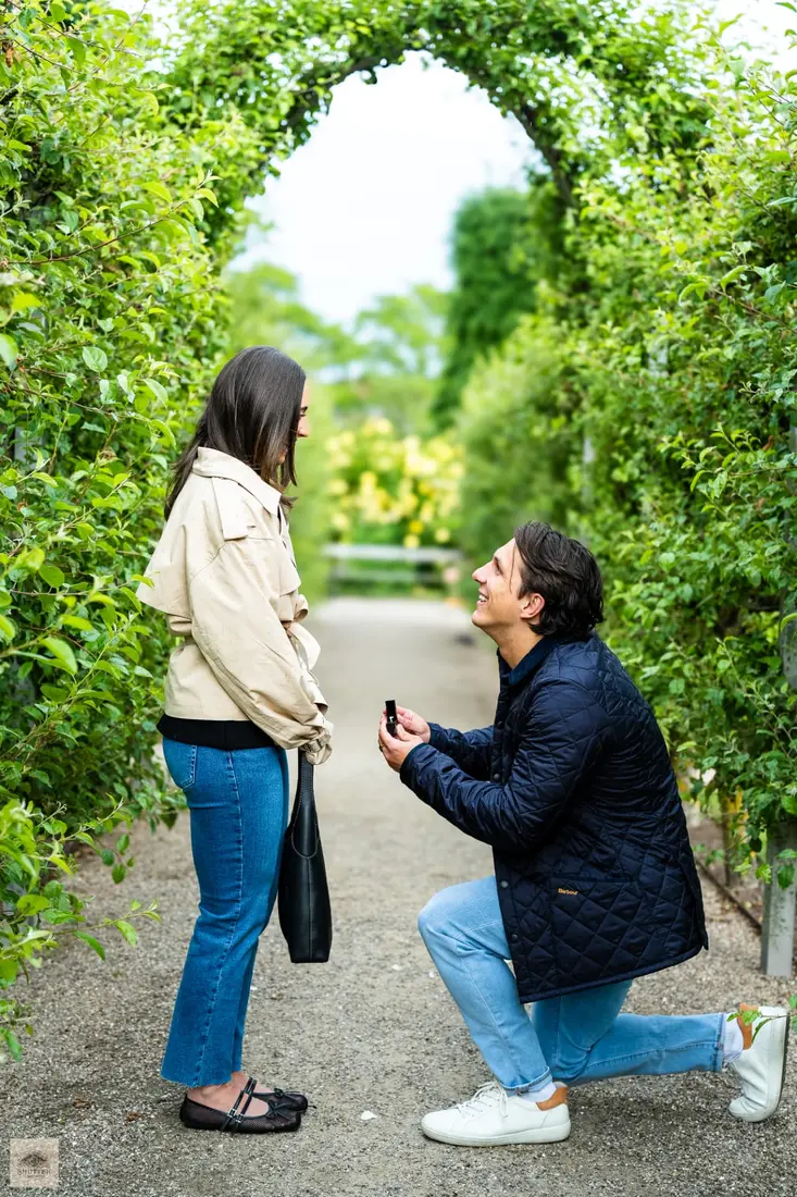 Surprise proposal photography Copenhagen — man kneeling with a ring under a green garden archway at Rosenborg